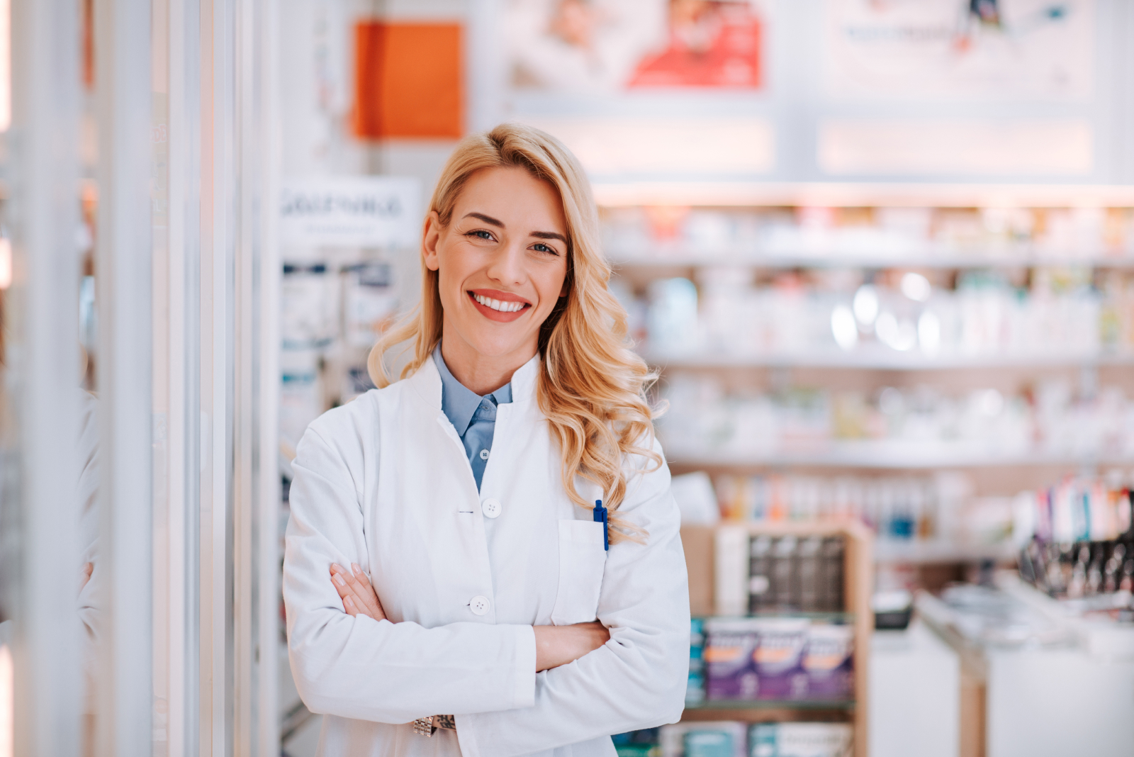 Eine lächelnde, blonde Frau im weißen Kittel steht mit verschränkten Armen vor einem Regal mit Medikamenten in einer Apotheke.